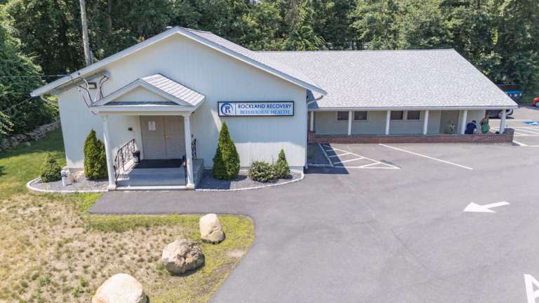 Front of the Rockland Recovery Behavioral Health building in Sharon, a grey single storey building with a pitched roof, columned entrance porch and the name board mounted on the gable