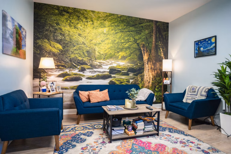 Therapy lounge with a blue sofa and two armchairs, a full wall mural of a woodland stream, table lamps and a low coffee table