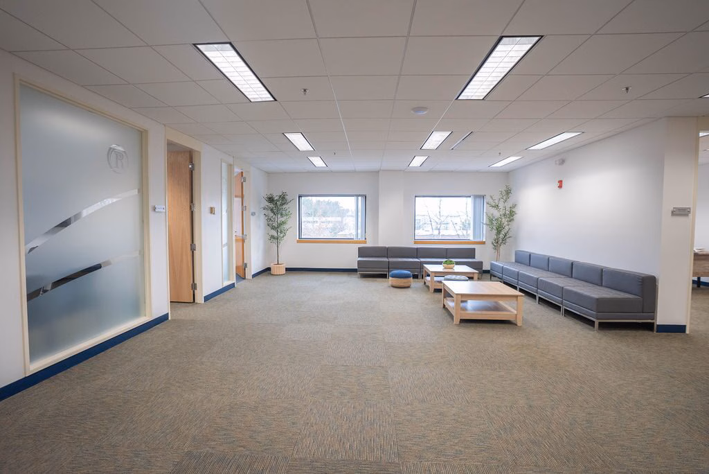 An open common area with long grey sofas, low wooden tables, potted trees and windows on two sides at Rockland Recovery Behavioral Health North in Bedford, Massachusetts.
