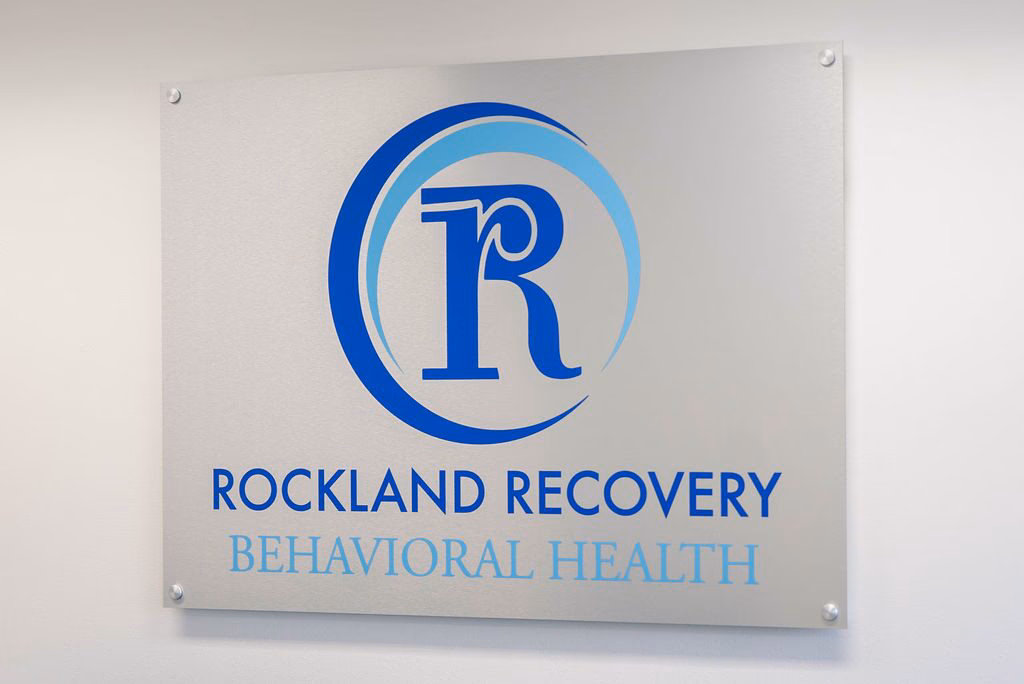 A brushed metal Rockland Recovery Behavioral Health sign mounted on a white wall.